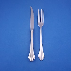 May include: A stainless steel knife and fork set against a blue background. The knife has a serrated blade and a decorative handle. The fork has four tines and a matching handle. The silverware is designed for formal dining.