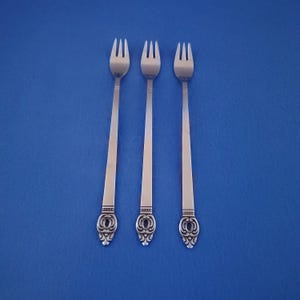May include: Three silver forks with a decorative design on the handles are displayed against a blue background. The forks have three tines and a detailed pattern near the base of the handle.