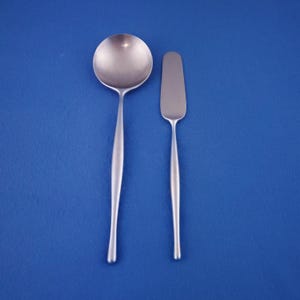 May include: A stainless steel soup spoon and butter knife set against a blue background. The spoon has a rounded bowl and a long, slender handle. The butter knife has a rectangular blade and a matching handle. The set is designed with a minimalist aesthetic.