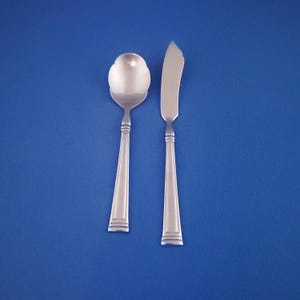 May include: A silver spoon with a flower-shaped bowl and a butter knife are displayed against a blue background. The spoon and knife have a simple, elegant design, with a tapered handle and decorative lines near the base.