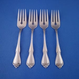 May include: Four silver forks with a floral design on the handle and a diamond-shaped cutout in the tines. The forks are arranged on a solid blue background, showcasing their elegant design.