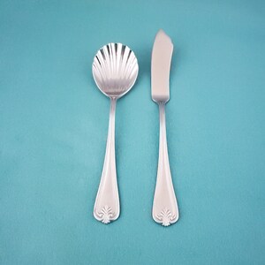 May include: A silver-toned metal spoon and a butter knife set. The spoon features a bowl shaped like a scalloped shell. Both utensils have a smooth, tapered handle that terminates in a decorative, embossed floral motif at the base of each piece.