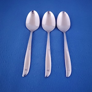 May include: Three matching silver-toned metal spoons arranged vertically. Each spoon features a smooth, polished bowl and a handle decorated with a subtle, etched starburst pattern near the base and a delicate, curved line running along the side.