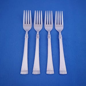 May include: Four silver forks with a classic design, arranged on a blue background. Each fork features a tined head, a detailed handle with vertical lines, and a decorative band near the top and bottom. The forks are evenly spaced.