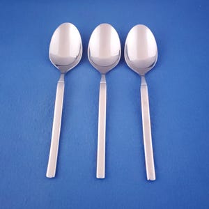 May include: Three stainless steel spoons with oval bowls and straight, rectangular handles are arranged on a blue background. The spoons are reflective, showing the light and the background. The spoons are for eating.