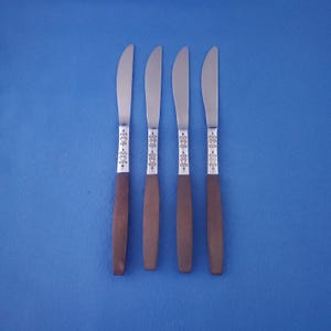 May include: Four butter knives with stainless steel blades and brown handles. The handles have a decorative silver design near the blade. The knives are arranged in a row against a blue background.