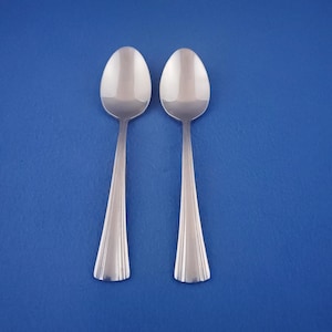 May include: Two stainless steel spoons with oval bowls and fluted handles are displayed against a solid blue background. The spoons are positioned vertically, side-by-side, reflecting light.