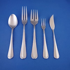 May include: A set of five stainless steel silverware pieces, including a spoon, two forks, a smaller fork, and a butter knife, arranged against a solid blue background. The silverware has a simple, modern design with tapered handles.