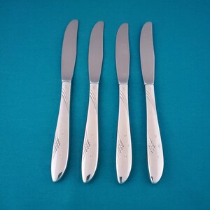 May include: A set of four silver-toned metal dinner knives arranged vertically. Each knife features a smooth, curved handle decorated with an engraved pattern consisting of three diagonal lines, a small cross-hatched grid, and several tiny star shapes.