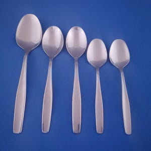 May include: Five stainless steel spoons of varying sizes are arranged against a solid blue background. The spoons have a simple, modern design with oval bowls and long, slender handles. The spoons are reflective, showing the light.