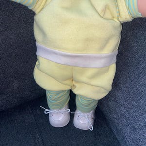 Op de afbeelding: Een pop draagt een lichtgeel shirt met korte mouwen, een witte tailleband en een bijpassende gele korte broek. Onder de broek draagt de pop een legging met dunne horizontale strepen in lichtblauw en geel. De pop draagt witte veterschoenen.