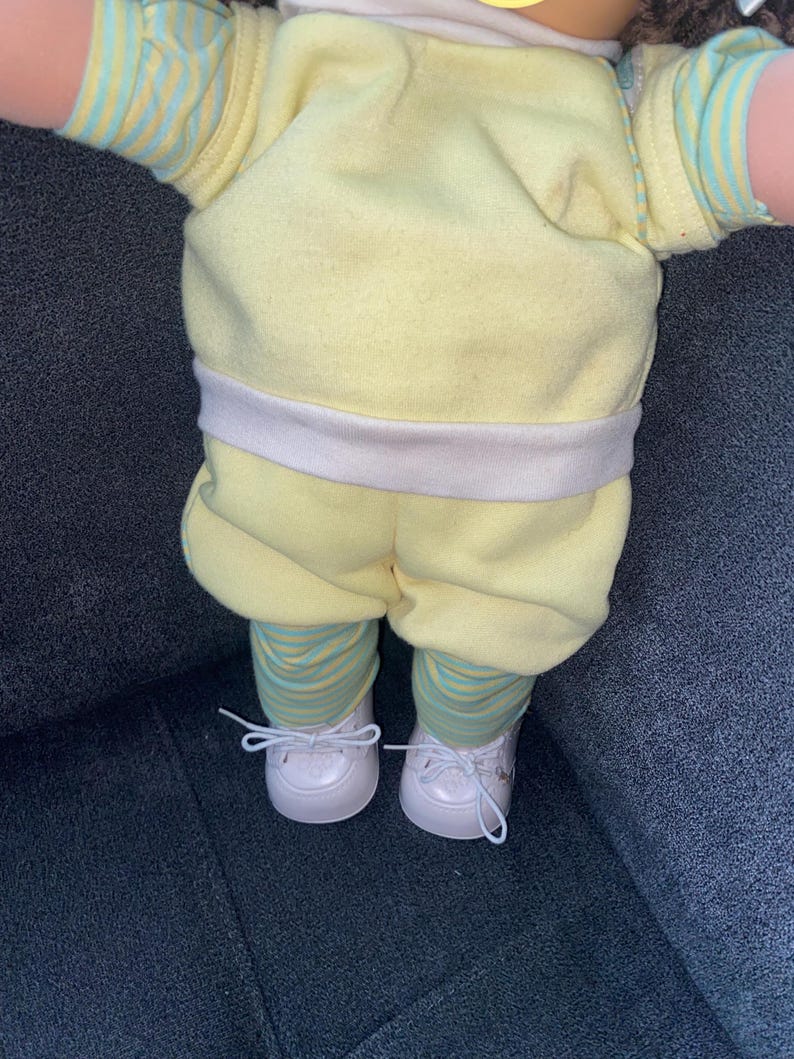 Op de afbeelding: Een pop draagt een lichtgeel shirt met korte mouwen, een witte tailleband en een bijpassende gele korte broek. Onder de broek draagt de pop een legging met dunne horizontale strepen in lichtblauw en geel. De pop draagt witte veterschoenen.