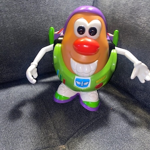 Puede incluir: Un juguete de Mr. Potato Head con el tema de Buzz Lightyear. El juguete presenta una cabeza en forma de patata de color canela con un casco morado, una nariz roja y una boca ancha y sonriente. Tiene detalles en verde y blanco, incluyendo una placa pectoral verde con el logo de Lightyear.