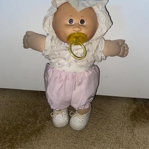 Peut inclure: Une poupée au corps souple avec un visage rond, des yeux marron et des cheveux en laine, portant un bonnet blanc à œillets, un haut blanc à motifs floraux, un bloomer rose clair et des chaussures blanches. Une tétine jaune est dans la bouche.