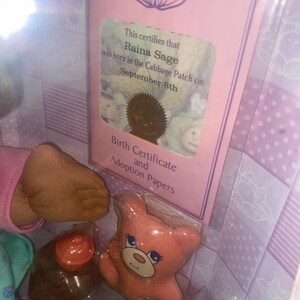 Pu&ograve; includere: Una confezione di bambola Cabbage Patch Kids con un certificato di nascita per Raina Sage nata l'8 settembre, un orsacchiotto di plastica rosa e un biberon marrone, su uno sfondo viola chiaro con un motivo a foglie di cavolo.