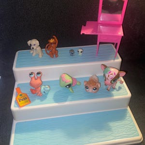 Puede incluir: Una colección de juguetes de Littlest Pet Shop, que incluye un perro blanco, un mono marrón y varias figuras de animales. También hay un tocador rosa con un espejo y la inscripción "Littlest Pet Shop". Los juguetes están dispuestos en una plataforma de varios niveles.