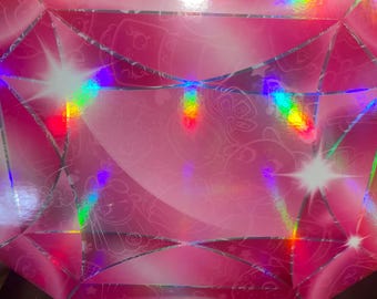 May include: A rectangular cardboard box with a vibrant pink, holographic finish that mimics the faceted appearance of a gemstone. The surface features a subtle, repeating line-art pattern of cartoon characters and stars, accented by bright light reflections.