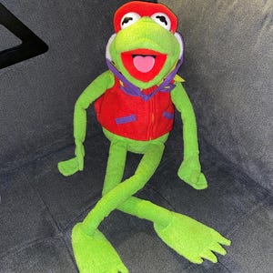 Pode incluir: Um peluche de sapo verde com um boné vermelho, um colete vermelho com detalhes roxos e um lenço roxo. O sapo tem olhos brancos grandes com pupilas pretas, uma boca bem aberta com uma língua rosa e braços e pernas longos e finos.