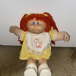 May include: A doll with bright orange yarn hair styled in pigtails, blue eyes, and a round face. The doll wears a yellow dress with white polka dots, a white bib featuring an embroidered giraffe, and white shoes with laces tied at the front.