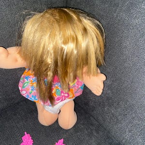 Puede incluir: Una mu&ntilde;eca rubia con un vestido rosa de flores y un pa&ntilde;al blanco. La mu&ntilde;eca est&aacute; sentada sobre una superficie gris. Hay dos pinzas para el pelo rosas con extensiones de pelo rubio en la superficie.