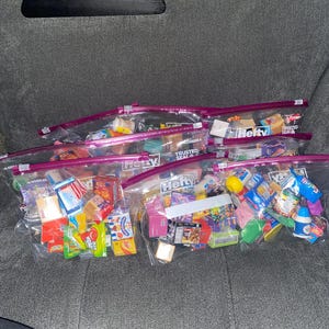 Puede incluir: Varias bolsas de plástico transparente con cierre de cremallera llenas de alimentos en miniatura. Las bolsas contienen una variedad de réplicas coloridas a pequeña escala de snacks, dulces y productos de supermercado. Las bolsas están dispuestas sobre una superficie gris.