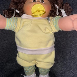 Op de afbeelding: Een pop met bruin wollen haar in staartjes met witte linten, een rond gezicht, bruine ogen en een gele fopspeen. De pop draagt een geel shirt met korte mouwen met lichtblauw gestreepte mouwen, een gele broek en witte schoenen.