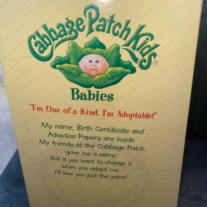 Pu&ograve; includere: Una scatola rettangolare di prodotto Cabbage Patch Kids Babies con sfondo giallo pallido e linee bianche diagonali. La scatola presenta un logo verde, un grafico di un bambino in una foglia di cavolo e il testo: I'm One of a Kind. I'm Adoptable!