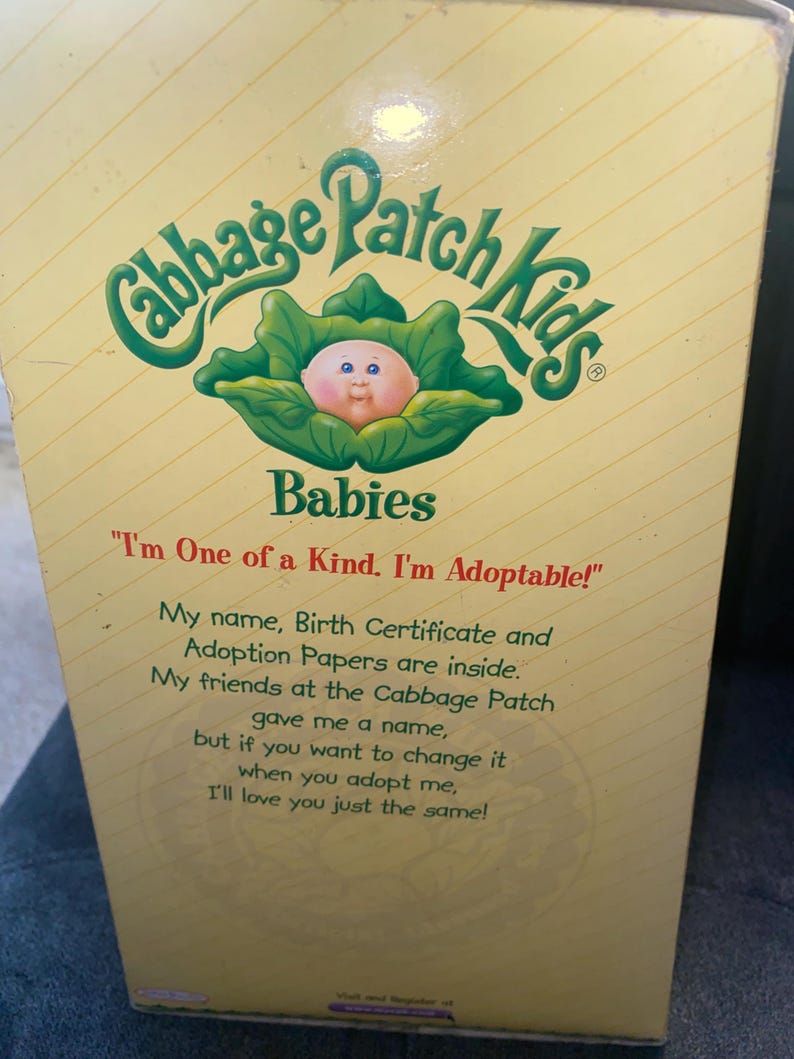 Pu&ograve; includere: Una scatola rettangolare di prodotto Cabbage Patch Kids Babies con sfondo giallo pallido e linee bianche diagonali. La scatola presenta un logo verde, un grafico di un bambino in una foglia di cavolo e il testo: I'm One of a Kind. I'm Adoptable!