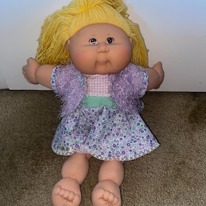 May include: A soft-bodied doll with yarn-like yellow hair, brown eyes, and a round face. The doll wears a purple fuzzy bolero jacket over a dress featuring a white and purple floral print, a light green waistband, and a white lace trim at the hem.