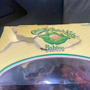 Pu&ograve; includere: Una scatola di cartone gialla Cabbage Patch Kids Babies con il lembo superiore danneggiato. La confezione presenta testo verde e un grafico di una testa di bambola all'interno di una foglia di cavolo. Una finestra in plastica rivela una bambola.
