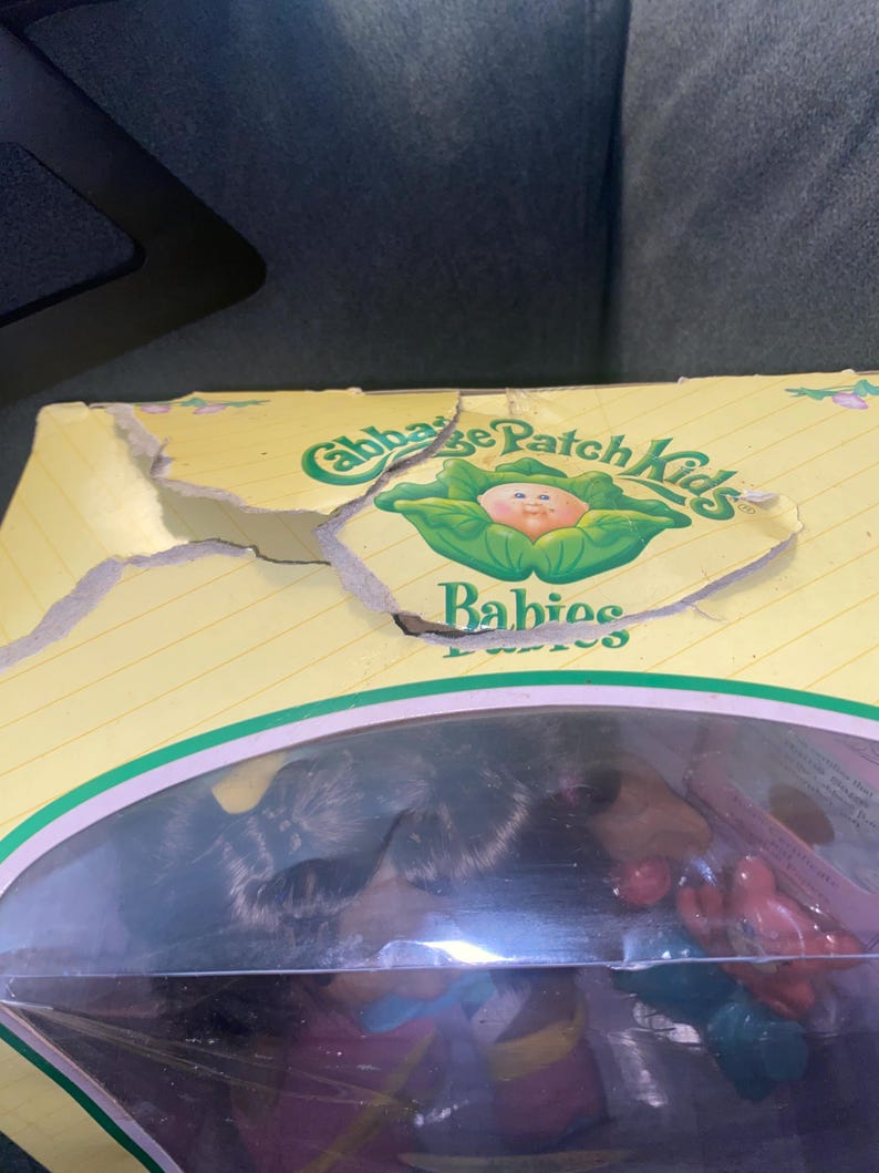 Pu&ograve; includere: Una scatola di cartone gialla Cabbage Patch Kids Babies con il lembo superiore danneggiato. La confezione presenta testo verde e un grafico di una testa di bambola all'interno di una foglia di cavolo. Una finestra in plastica rivela una bambola.