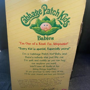 Pu&ograve; includere: Una scatola di cartone rettangolare per una bambola Cabbage Patch Kids Babies. La confezione gialla presenta il logo verde, un grafico di un bambino in una foglia di cavolo e testo stampato che include le frasi I'm One of a Kind e I'm Adoptable.