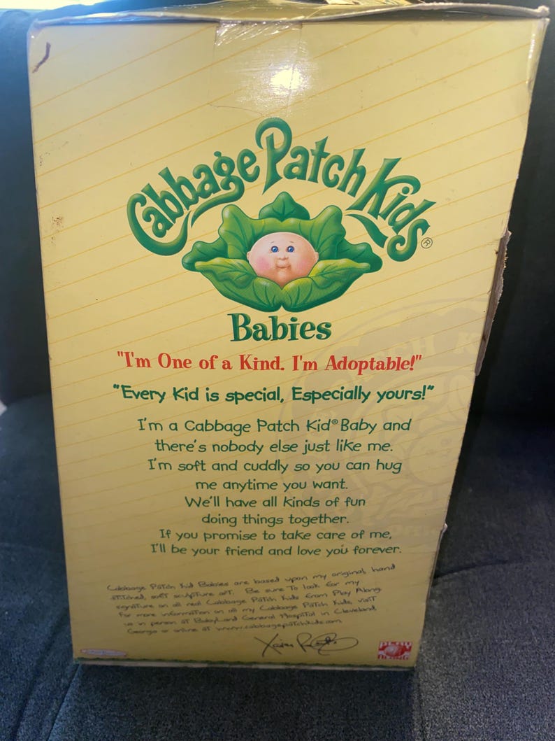 Pu&ograve; includere: Una scatola di cartone rettangolare per una bambola Cabbage Patch Kids Babies. La confezione gialla presenta il logo verde, un grafico di un bambino in una foglia di cavolo e testo stampato che include le frasi I'm One of a Kind e I'm Adoptable.