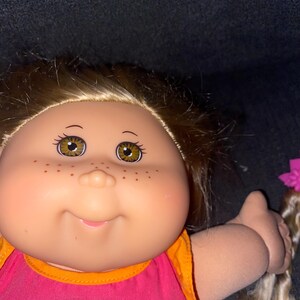 Puede incluir: Una mu&ntilde;eca Cabbage Patch Kids con cabello rubio, ojos marrones y pecas. Lleva un vestido rosa y naranja con estampado floral y el texto "CPK" en la parte delantera.