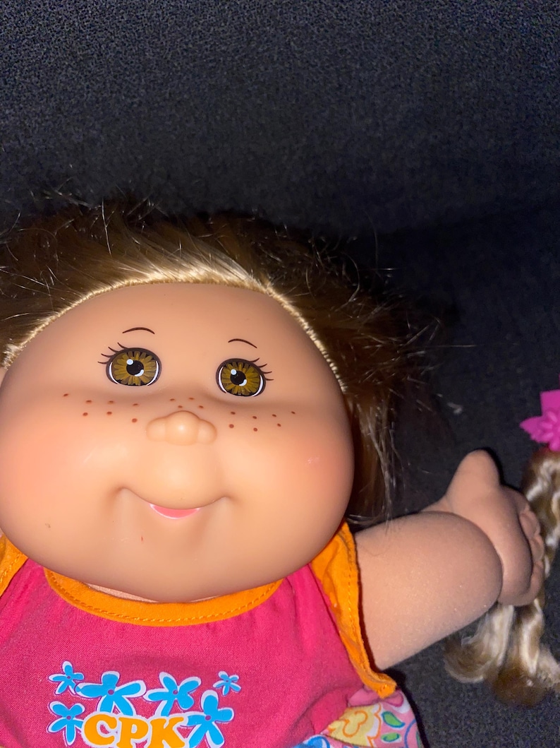 Puede incluir: Una mu&ntilde;eca Cabbage Patch Kids con cabello rubio, ojos marrones y pecas. Lleva un vestido rosa y naranja con estampado floral y el texto "CPK" en la parte delantera.