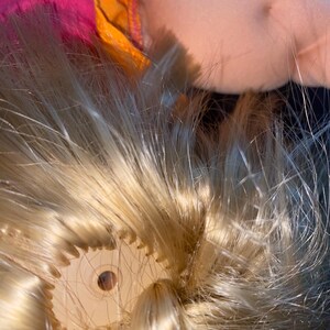 Puede incluir: Primer plano de la cabeza de una mu&ntilde;eca con cabello rubio. El cabello est&aacute; recogido hacia atr&aacute;s y asegurado con una liga de pl&aacute;stico. La liga es visible a trav&eacute;s del cabello.