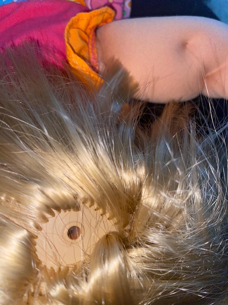 Puede incluir: Primer plano de la cabeza de una mu&ntilde;eca con cabello rubio. El cabello est&aacute; recogido hacia atr&aacute;s y asegurado con una liga de pl&aacute;stico. La liga es visible a trav&eacute;s del cabello.