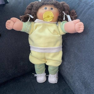 Pode incluir: Uma boneca com cabelo de lã castanho em tranças com fitas brancas, com rosto redondo, olhos azuis e uma chupeta amarela. A boneca veste uma blusa de manga curta amarelo-pálido com cintura branca, leggings às riscas verde-claras e amarelas e sapatos brancos.