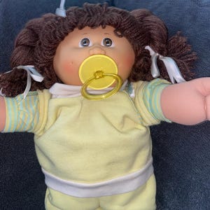 Op de afbeelding: Een pop met bruin garen haar in staartjes met witte linten, met een rond gezicht, roze wangen en bruine ogen. De pop draagt een geel shirt met korte mouwen met lichtblauwe strepen en heeft een gele fopspeen in de mond.