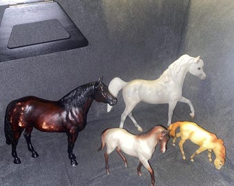 May include: Four toy horse figurines in various colors. One is a dark brown with black mane and tail, another is white with gray spots, and two are tan and brown. The horses are posed on a gray surface.