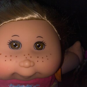 Puede incluir: Primer plano de una mu&ntilde;eca Cabbage Patch Kid con cabello rubio, ojos marrones y pecas. La mu&ntilde;eca lleva un vestido rosa y naranja con estampado floral y el texto "CPK" en la parte delantera.