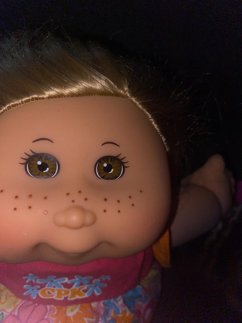 Puede incluir: Primer plano de una mu&ntilde;eca Cabbage Patch Kid con cabello rubio, ojos marrones y pecas. La mu&ntilde;eca lleva un vestido rosa y naranja con estampado floral y el texto "CPK" en la parte delantera.