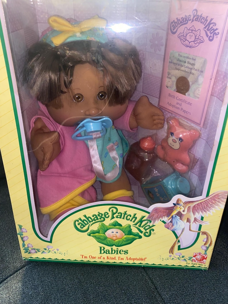 Pu&ograve; includere: Una bambola Cabbage Patch Kids con capelli e occhi castani, che indossa un completo rosa e giallo con un ciuccio blu. La bambola &egrave; in una scatola con un certificato di nascita, un piccolo orso rosa e un biberon. La scatola mostra una cicogna.
