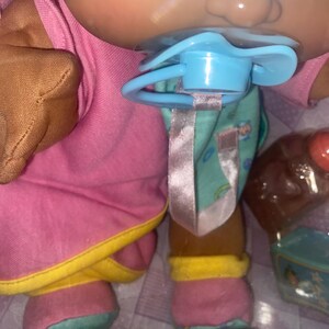Pu&ograve; includere: Una bambola Cabbage Patch Kids dalla carnagione abbronzata, che indossa un vestito rosa con bordi gialli e scarpe rosa e verde acqua. La bambola ha un ciuccio azzurro in bocca attaccato a un nastro rosa ed &egrave; confezionata con un piccolo biberon marrone.