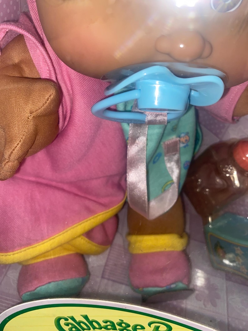 Pu&ograve; includere: Una bambola Cabbage Patch Kids dalla carnagione abbronzata, che indossa un vestito rosa con bordi gialli e scarpe rosa e verde acqua. La bambola ha un ciuccio azzurro in bocca attaccato a un nastro rosa ed &egrave; confezionata con un piccolo biberon marrone.