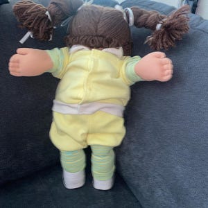 Op de afbeelding: Een pop met bruin wollen haar in twee staartjes vastgebonden met witte linten. De pop draagt een lichtgeel shirt met lange mouwen en een witte kraag, een bijpassende gele broek en een lichtgroene en geel gestreepte legging met witte schoenen.