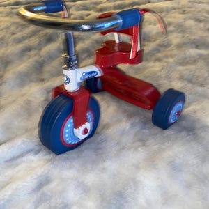 May include: A red and blue tricycle with a chrome handlebar. The tricycle has blue wheels with white stars and red accents. The brand name "Bitty Baby" is visible on the front.