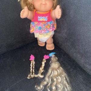 Może przedstawiać: Lalka Cabbage Patch Kids z blond włosami, ubrana w różową sukienkę w kwiaty i pieluchę. Lalka trzyma ramiona wyciągnięte. Na ciemnoszarym tle leżą trzy blond pasma włosów z różowymi spinkami.