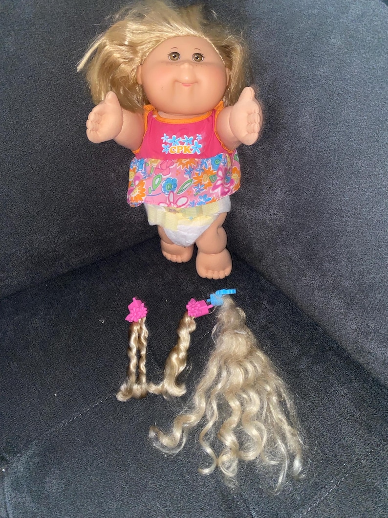 Puede incluir: Una mu&ntilde;eca Cabbage Patch Kids con cabello rubio que lleva un vestido rosa con estampado floral y un pa&ntilde;al. La mu&ntilde;eca tiene los brazos extendidos. Hay tres mechones de cabello rubio con pinzas rosas sobre una superficie gris oscuro.
