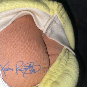 Op de afbeelding: Een pop met een huid van beige stof die een lichtgeel kledingstuk draagt. De pop heeft een blauwe inkt-handtekening op de onderrug met de tekst Xavier Roberts '85. De stof van het kledingstuk is gestructureerd en lichtgeel van kleur.